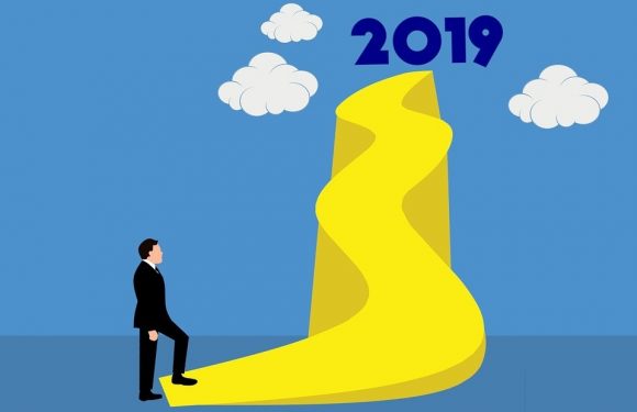 A man in a suit looking up at a yellow twisted road leading to the year 2019, with white clouds and a blue sky in the background.