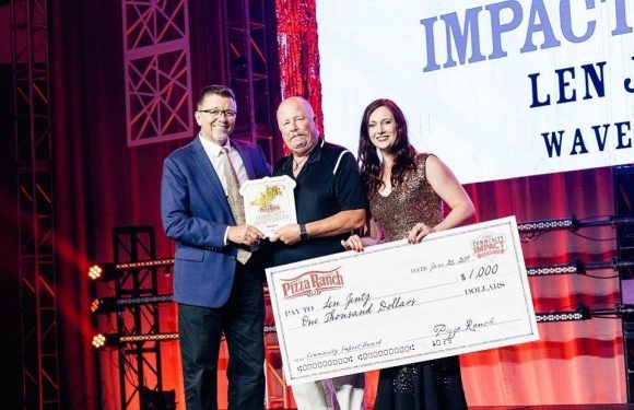 Three people stand on a stage holding a large check for $1,000 made out to Len Genty and a plaque. The backdrop displays the words "IMPACT" and "LEN" with a red and white design.