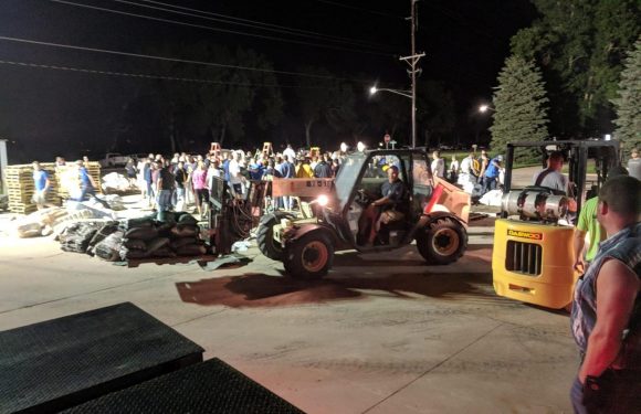 A forklift is moving materials near a crowd of people outside at night, with some individuals wearing uniforms and several safety cones and barriers present.