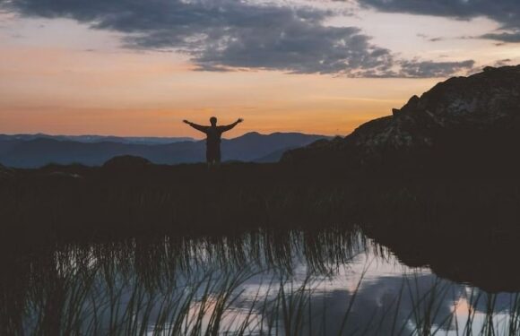 A person stands on a ridge with arms outstretched at sunset, overlooking mountains, with a body of water reflecting the sky and surrounding landscape.