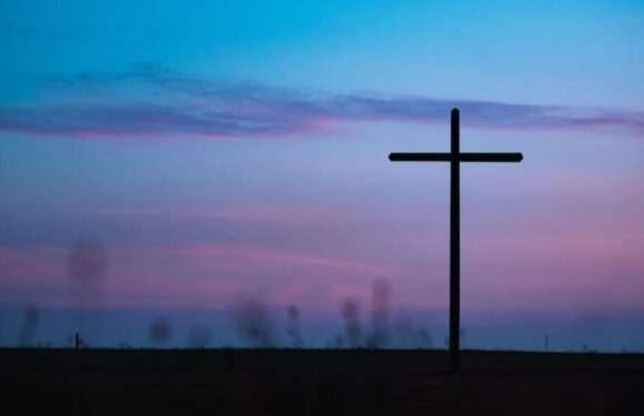 A large cross silhouette stands against a colorful evening sky with pink and blue hues, and a flat, dark landscape in the foreground.