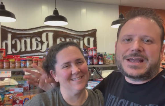 A man and woman smiling and posing for a selfie inside a restaurant with a sign in the background that says "Pizza Ranch." They are standing near shelves of food products.