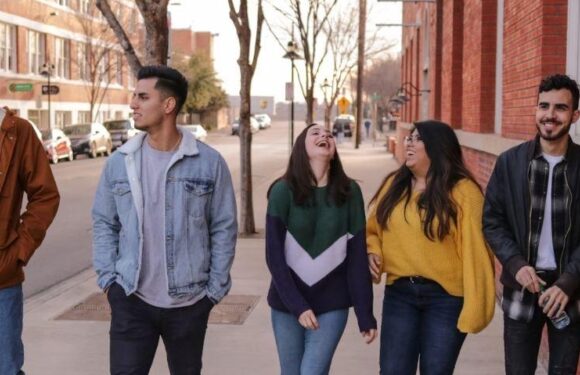Five young adults walk along a sidewalk during daytime, laughing and chatting together, with brick buildings and trees in the background.