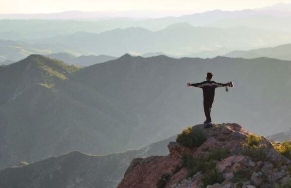 A person in a Pizza Ranch uniform stands on a rocky cliff with arms outstretched, overlooking a vast mountain range during daylight.
