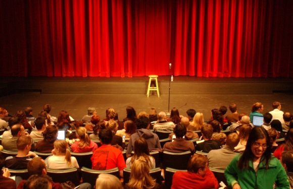 A stage with a red curtain, a stool, and a microphone stand in front of an audience seated facing the stage, some using laptops or phones.
