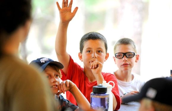 A young boy in a red shirt with short dark hair raises his hand while sitting at a table with other children. The children are indoors with a blurred background.
