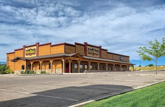 A large, empty parking lot in front of a Pizza Ranch restaurant with a wooden exterior, green grass, and a partly cloudy sky overhead.