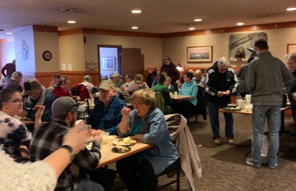 A group of people dining in a crowded restaurant, some wearing uniforms with name tags, with a mix of older and younger individuals enjoying their meal.