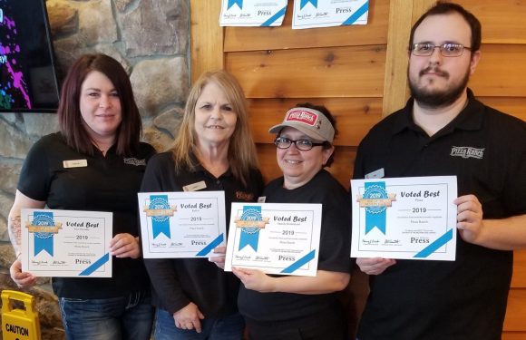 Four employees in black shirts holding certificates stand inside a wood-paneled room with a stone wall. They are smiling, and the certificates say "Voted Best 2019" and "Press."