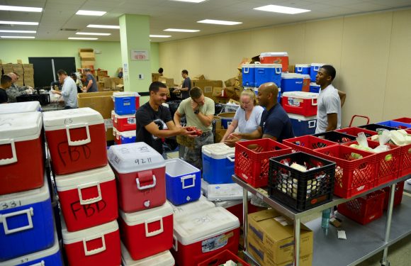 People in uniform working together in a large room filled with packed coolers, boxes, and supplies for a community or charity event, possibly at Pizza Ranch.