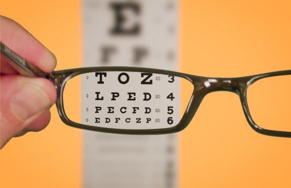 A hand holds a pair of glasses in front of an eye chart on a orange background. The glasses lens magnifies some of the letters on the chart.