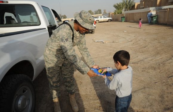 A soldier in camouflage uniform hands an item to a young boy in a gray sweatshirt outdoors. Other vehicles and people are visible in the background on a dirt lot.
