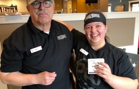 A man and a young woman pose indoors, both wearing black Pizza Ranch shirts. The man wears glasses and a gray cap, while the woman holds a small card and smiles.