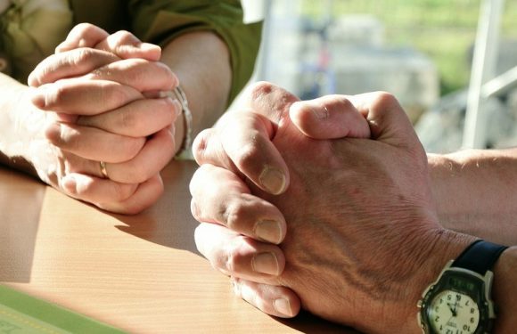Two people hold hands in a supportive manner, with one person's hands resting over the other's, on a table near a window with a blurred outdoor scene.
