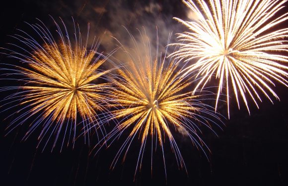 Three large fireworks burst in the night sky, with golden and violet streaks radiating outward against a dark background.