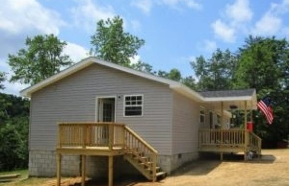 A small tan house with a wooden porch, stairs, and railing, surrounded by green trees and a clear blue sky. An American flag is on the porch.