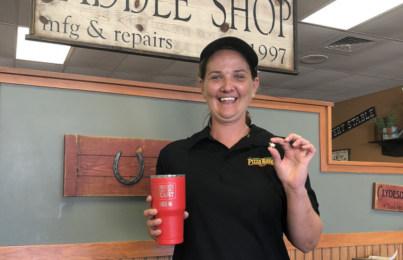 A smiling woman in a black Pizza Ranch uniform holding a red tumbler with a straw, standing inside a restaurant with wooden decor and signs.