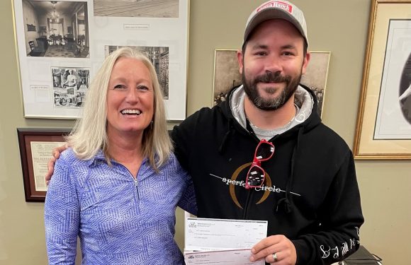 A woman and a man are smiling and standing close together indoors, holding a check and a piece of paper, with framed photos on the wall behind them.