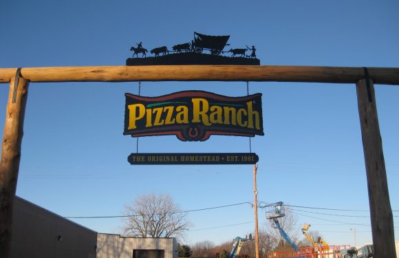 Sign hanging from a wooden arch with a silhouette wagon and oxen on top, reading "Pizza Ranch" and "The original homestead, est. 1981" with construction equipment in background.