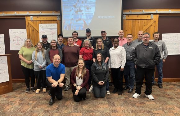 Group of people standing and kneeling indoors in front of a wooden wall, with presentation slides and charts on the sides, celebrating an event called "Ranchuation."