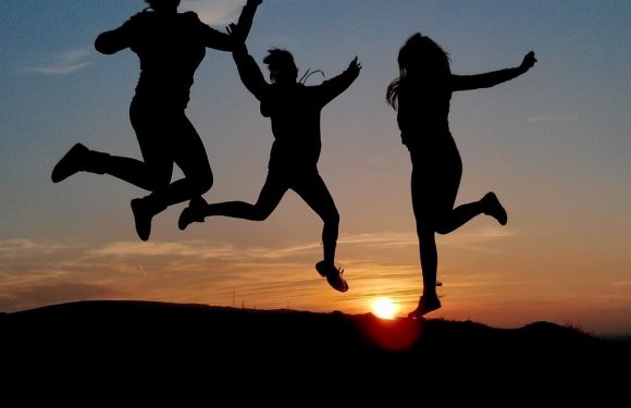 Three people are jumping and holding hands against a sunset sky, with a mixture of clouds and clear sky, silhouetted on a hill.