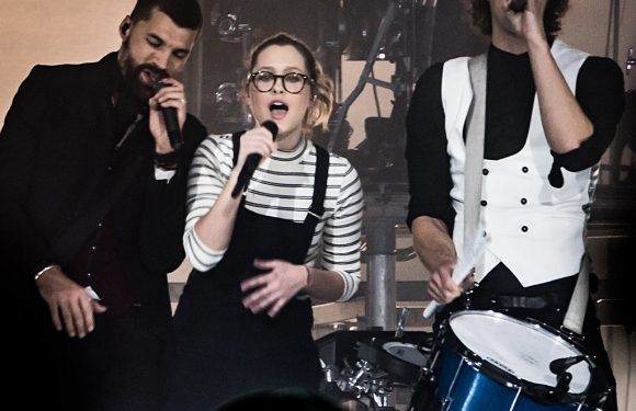 Three performers singing into microphones on stage, with a drummer partially visible behind them, in a dimly lit setting.
