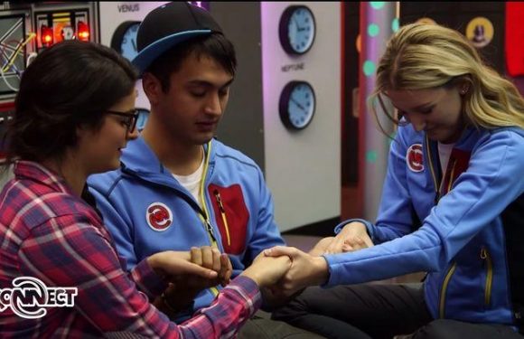 Three young women and one young man sit in a room with colorful decor, holding hands and forming a circle, appearing to share an emotional moment.