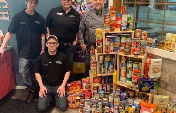 Four employees in black uniforms with name tags stand and kneel next to a large pyramid of canned and boxed food items at a restaurant, near a window.