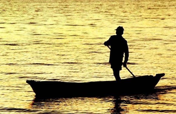 A silhouette of a person standing on a small boat holding a paddle, with a golden sunset reflection on calm water in the background.