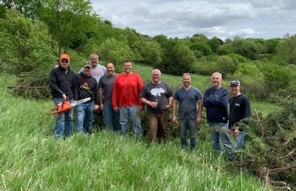 A group of nine men standing outdoors on a grassy area near a stream, with green trees and cloudy sky in the background. One man is holding a chainsaw.