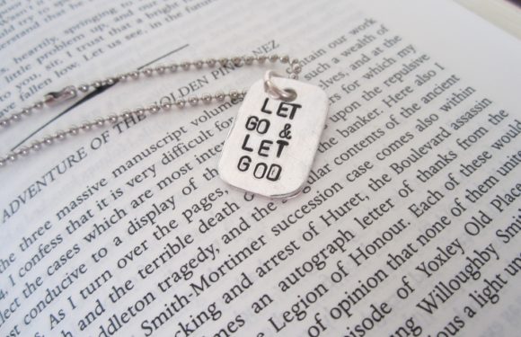 A metal dog tag with the words "LET & LET GO" on it, attached to a ball chain, resting on an open book with printed text.