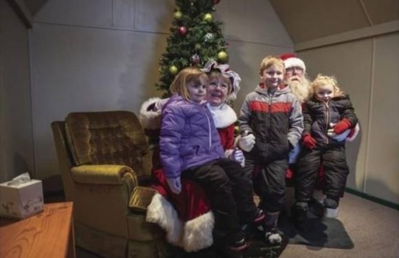 Four children in winter clothing are sitting on Santa's lap with a Christmas tree in the background, inside a cozy room decorated for the holidays.