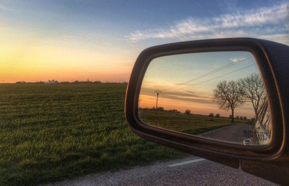 A sunset scene viewed through a car's side mirror, showing a road, leafless trees, power lines, and a colorful sky with orange, pink, and blue hues.