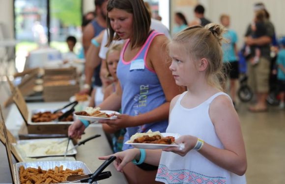 Two young girls, one with blonde hair in a bun wearing a white sleeveless top, and the other with shoulder-length brown hair in a blue tank top, hold plates of food at a buffet.