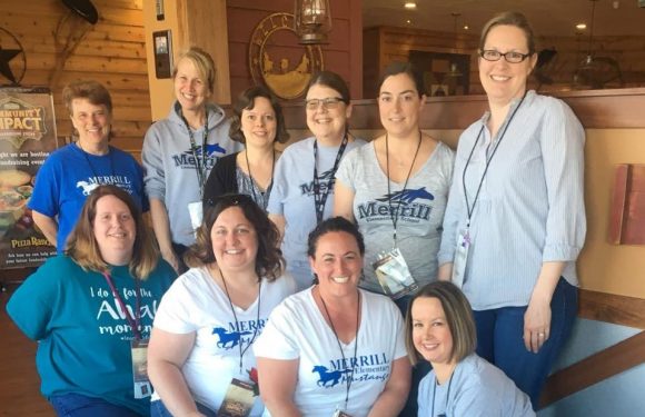 Group of nine women, some wearing Merrill Elementary School t-shirts or sweatshirts, smiling indoors with wooden walls and a framed picture in the background.