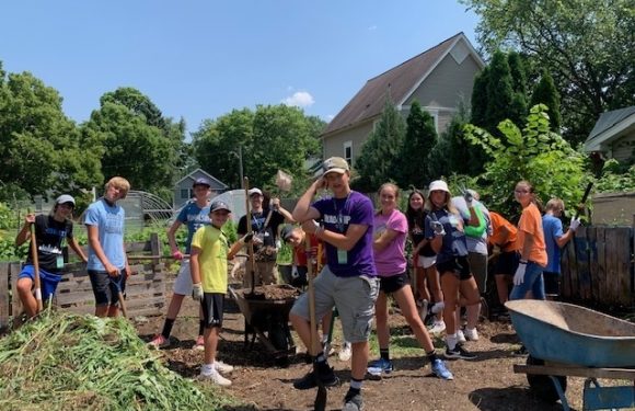 Group of children and teenagers working together in a garden, some holding shovels, with trees, a house, and blue sky in the background.