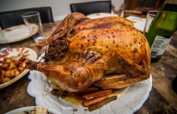 A golden-brown roasted turkey with crispy skin rests on a white serving platter, surrounded by a dinner table with plates, glasses, and green beverage bottles.