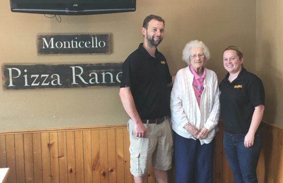 A woman with white curly hair and glasses standing between two young staff members in black shirts, inside a restaurant with wooden paneling and signs.