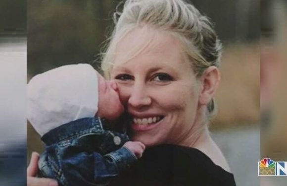 A woman smiling while holding a baby with a painted face, outdoors with a blurred background. The woman wears black, and the baby wears a white hat and denim jacket.