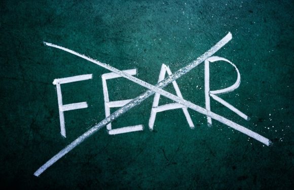 "The word 'FEAR' is written on a dark green surface with white chalk, crossed out with a large diagonal line, symbolizing overcoming fear."