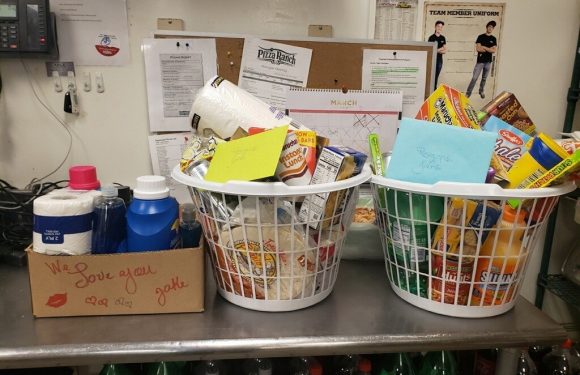 Two white baskets filled with snacks, notes, and cards sit on a metal counter next to a cardboard box with toiletries and a handwritten message.