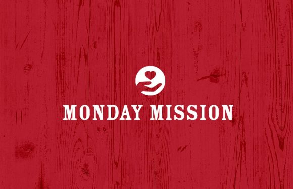 A red wooden background with a white logo of a smiling face holding a heart, and the text "MONDAY MISSION" in bold white letters.