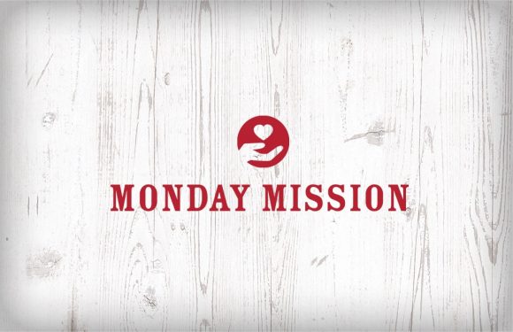 A white wooden background with a red circular logo featuring a hand holding a heart, and bold red text that reads "Monday Mission".