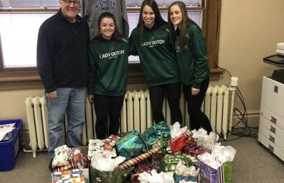 Four people, three young women in green "Lady Dutch" hoodies and one man in a black jacket, stand behind a table filled with wrapped Christmas gifts in an office.