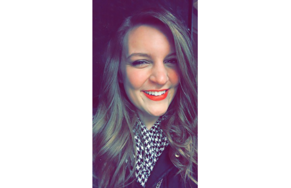 A woman with long, wavy, light brown hair smiling and winking, wearing bright red lipstick, a black jacket, and a patterned shirt.