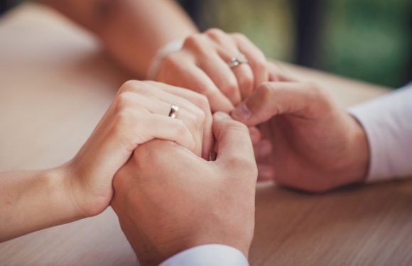 Two people are holding hands, with one person wearing a ring on the left hand. They are touching gently on a wooden table, with a blurred background.