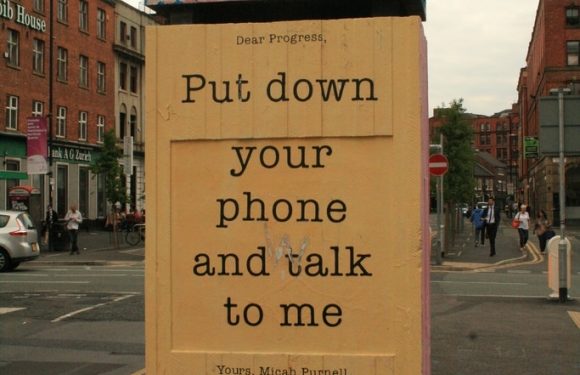 A large outdoor sign with a yellow background displays a message: "Dear Progress, put down your phone and talk to me," attributed to Micah Purnell, on a city street.