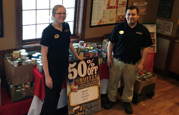 Two employees in black Pizza Ranch shirts stand next to tables of canned food for a charity drive, with a large "50% OFF Buffets with two cans of food" sign in front.