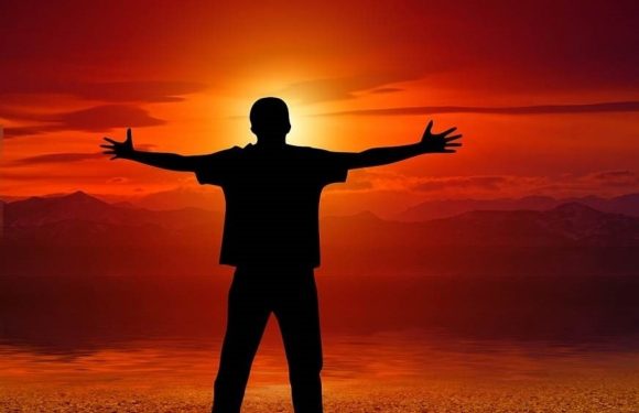 A person with outstretched arms standing by a body of water at sunset, with mountains in the distance and a warm, glowing sky.