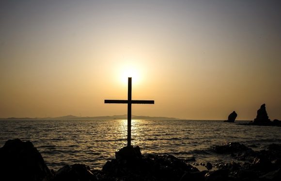 A cross on rocks near the water during sunset or sunrise with the sun shining behind, creating a silhouette against a calm sea and distant land.